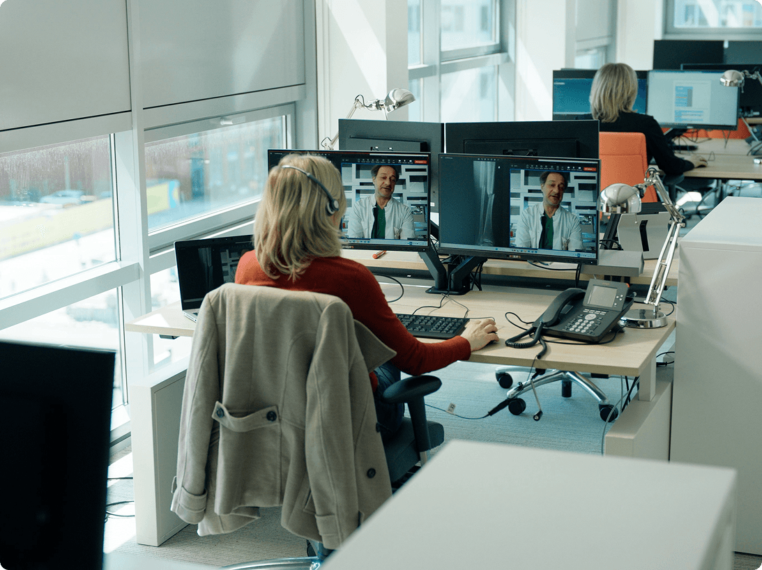 An image of a blond person with a headset sitting behind a dual monitor desk videocalling with another person.